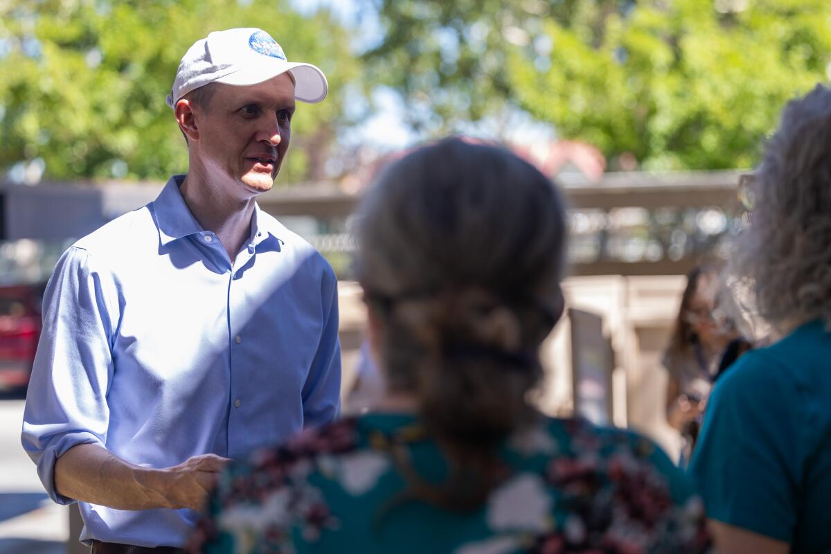 George Whitesides standing in the sun wearing a baseball cap and speaking to two people, their backs to the camera