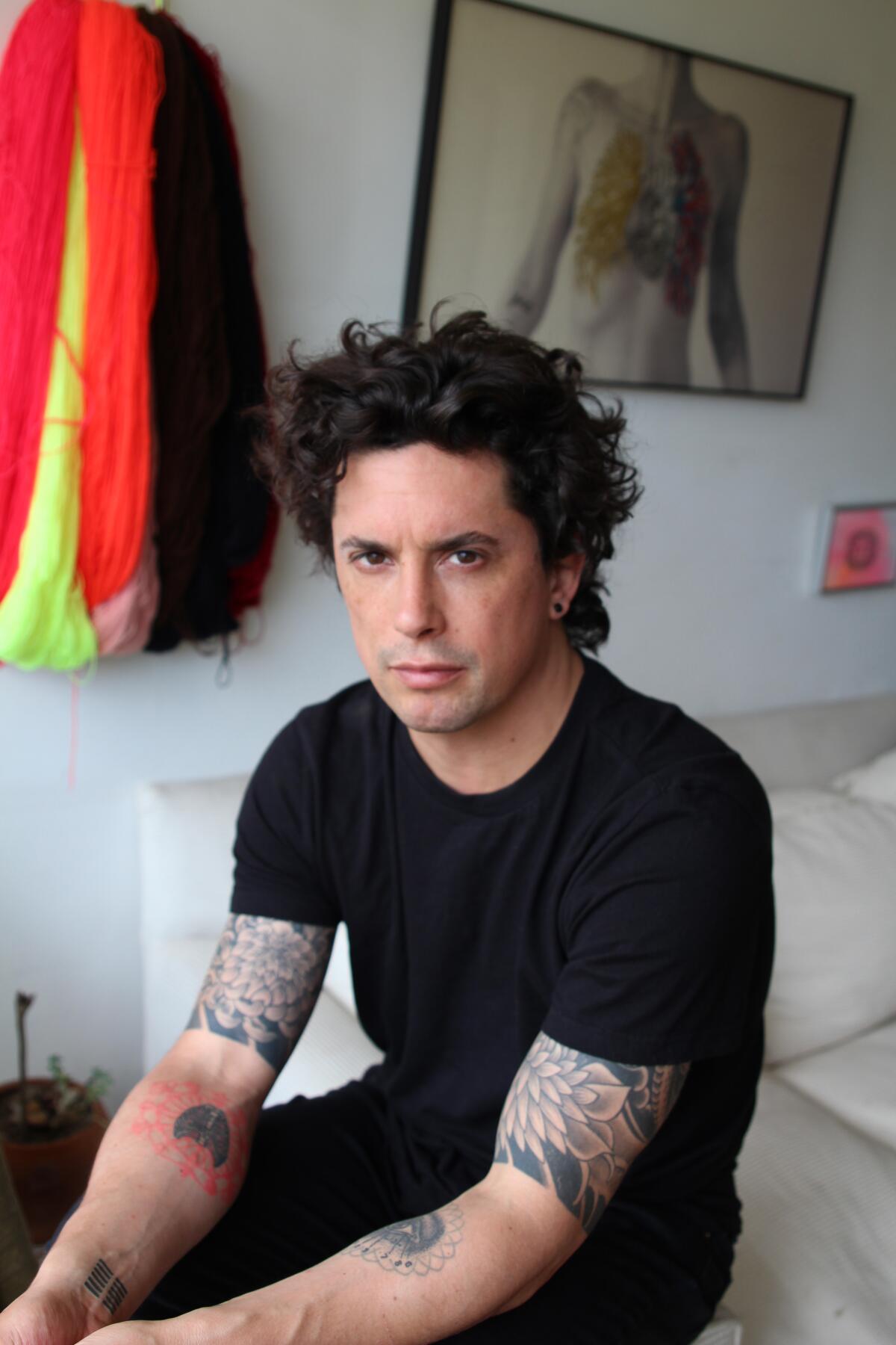 Benjamin Labatut, in black, is seen sitting on the arm of a white couch with a bright textile work hanging behind him