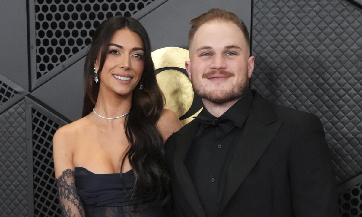 Brianna LaPaglia and Zach Bryan smiling in black formal attire in front of a black background with a gold logo