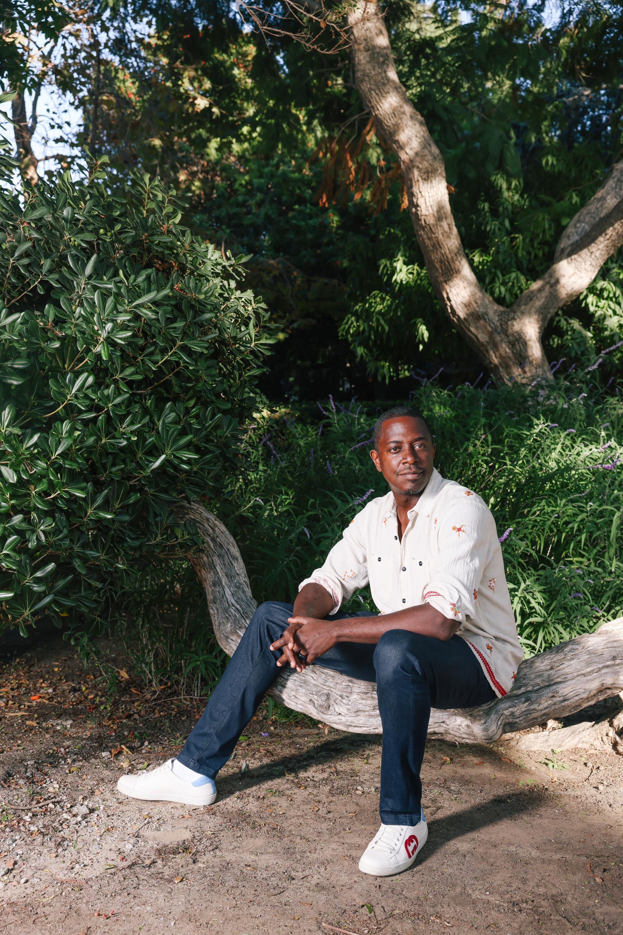 Sanford Biggers sits on a low tree branch in a cream-colored shirt and dark blue jeans