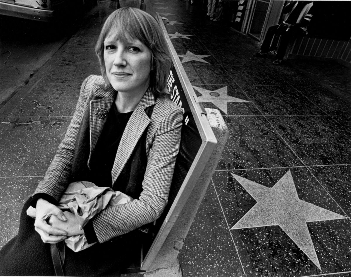 Eve Babitz sits on a bench near a sidewalk with stars on it in 1980.