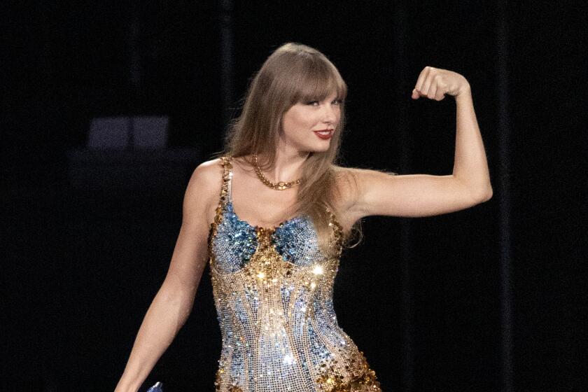 Taylor Swift in a bejeweled blue and gold leotard flexing her left arm and holding a microphone in her right hand on a stage