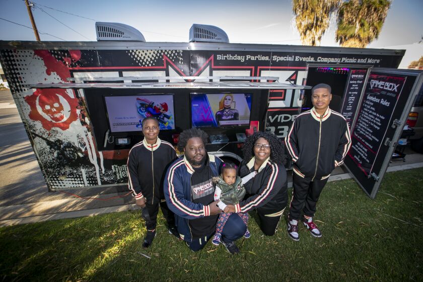 NORWALK, CA - NOVEMBER 10: Ebony Lynk, 38, and her husband Romel Mallard, 42, who are co-owners of Gamebox Mobile Video Gaming, and their kids: Romari Mallard, left, 10, Rori Mallard, 1, baby mascot, and Bryce Hayes, right, 13, game coaches, who help coach customers. Lynk and Mallard are among tens of thousands of California sole proprietors who have suffered severe losses from the COVID-19 pandemic. From their home in Paramount, they rent out a 20-foot trailer full of video games for parties `and fundraisers. They bought the business in November 2019, a few weeks before her husband was laid off from his commercial trucking job, and were just beginning to turn a profit when COVID-19 hit and customers vanished. "We went to zero revenue," she said. They applied for a disaster loan from the federal Small Business Administration, but like many micro-businesses, did not qualify. Then Lynk learned about the city/county $100 million Los Angeles Regional COVID-19 Recovery Fund. In October, she got a grant for $5000. "It made a huge difference," she said. "We were paying out of pocket for masks and gloves and supplies." And now that customers are trickling back, the grant also allows her to offer the trailer free of charge for occasional charity events at schools and nonprofits. Photos taken Tuesday, Nov. 10, 2020 in Norwalk, CA. (Allen J. Schaben / Los Angeles Times)