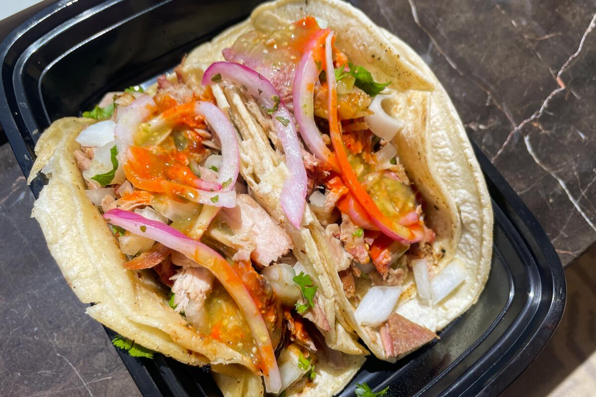Two carnitas tacos with onion, carrot and other garnish in a black plastic to-go container