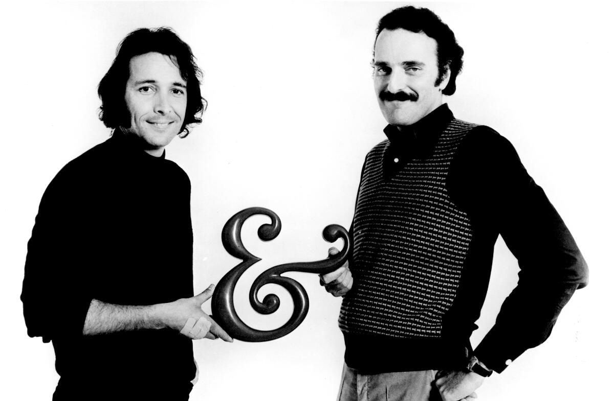 Herb Alpert and Jerry Moss, both in black shirts, hold a large ampersand between them.