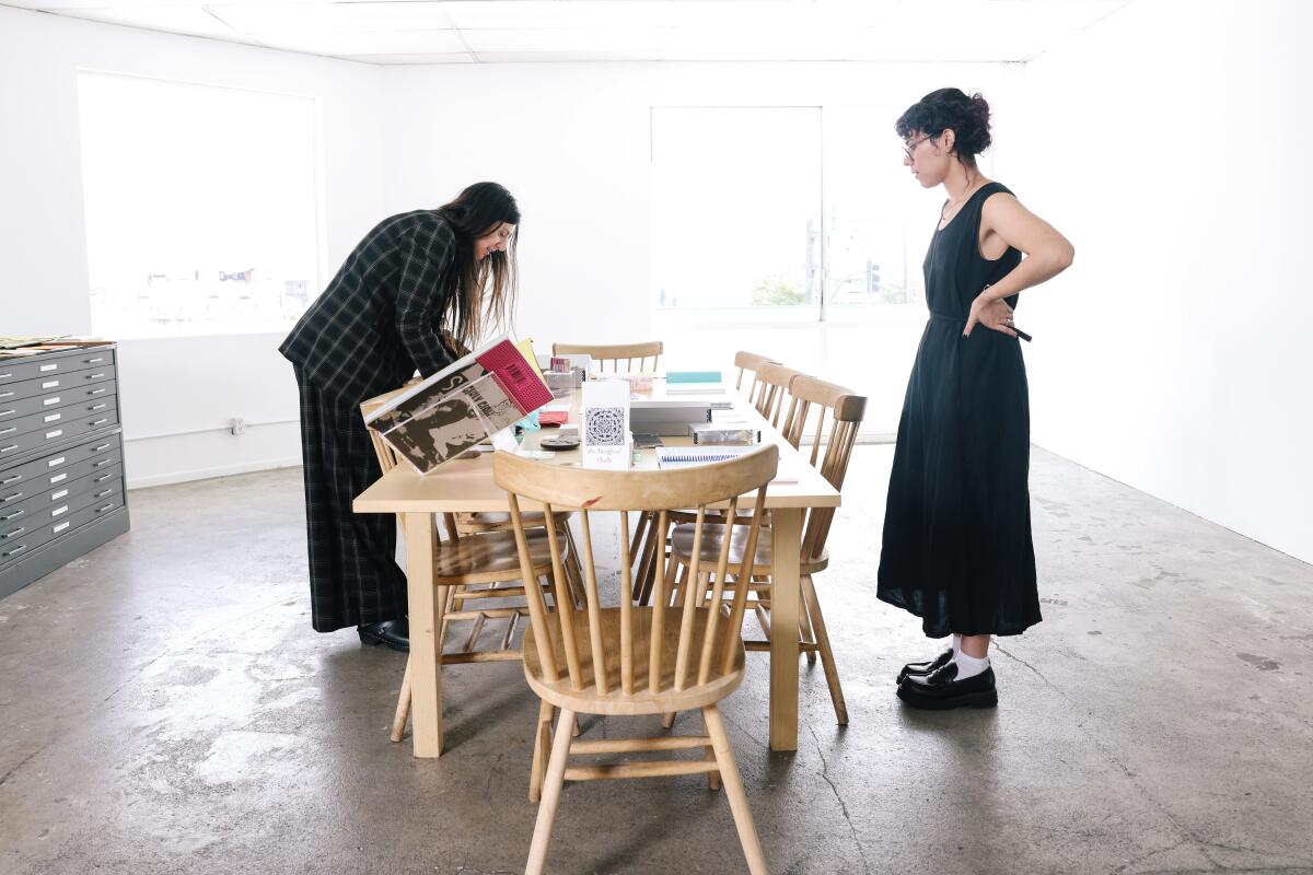 Two women in black on opposite sides of a wooden table with chairs that is full of artists' materials.