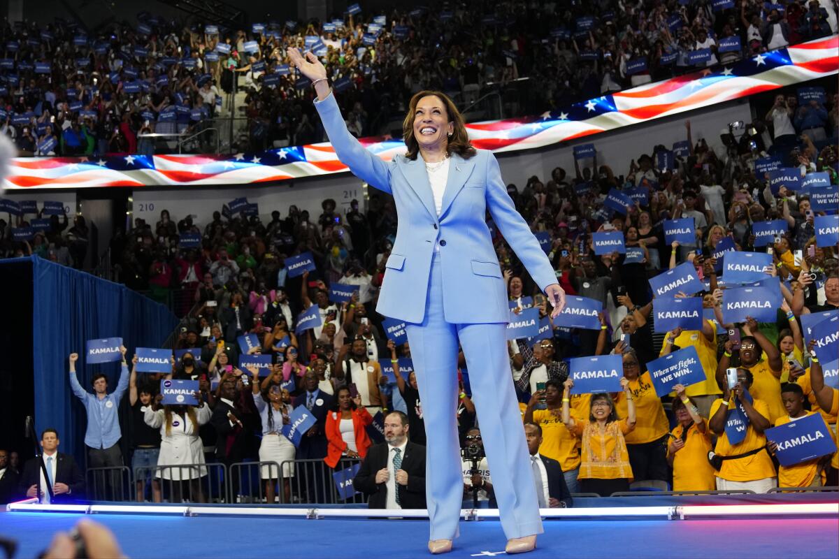 Kamala Harris waves to a large crowd, flag streamers and people holding "Kamala" signs behind her