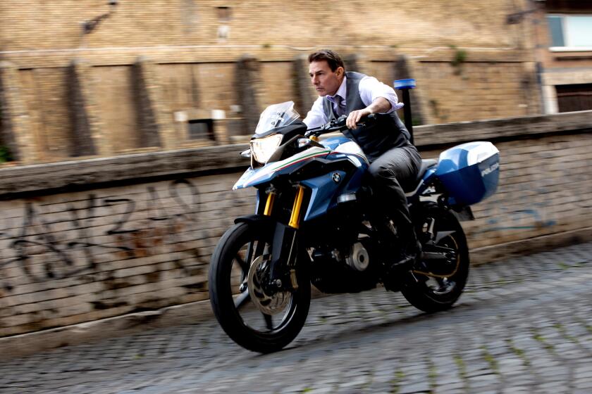 Tom Cruise wears a gray suit while riding a motorcycle down a cobblestone road.