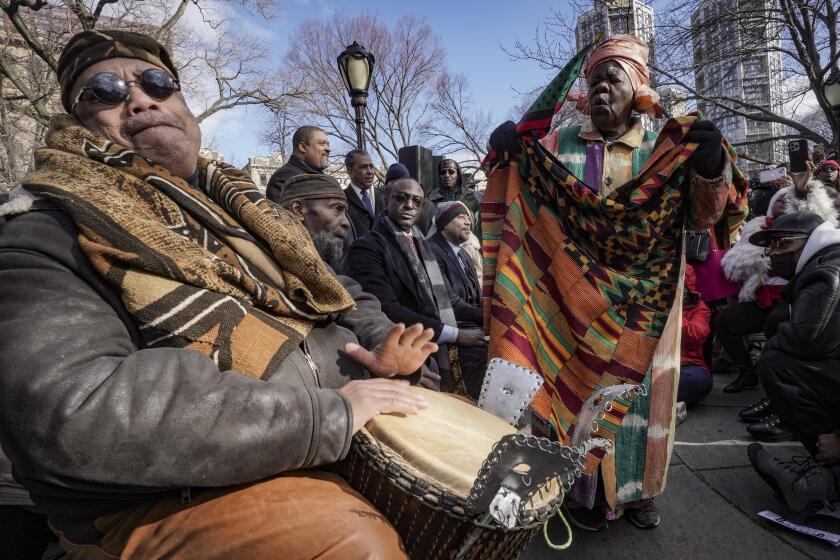 Master drummer Baba Don Babatunde, far left, and civil rights activist Queen Mother Moore, far right, lead a spiritual moment, during the Central Park gate-naming ceremony to honor the five men exonerated after being wrongfully convicted as teenagers for the 1989 rape of a jogger in Central Park, including Yusef Salaam, seated third from left, Raymond Santana Jr., seated fourth from left, Monday Dec. 19, 2022, in New York. (AP Photo/Bebeto Matthews)