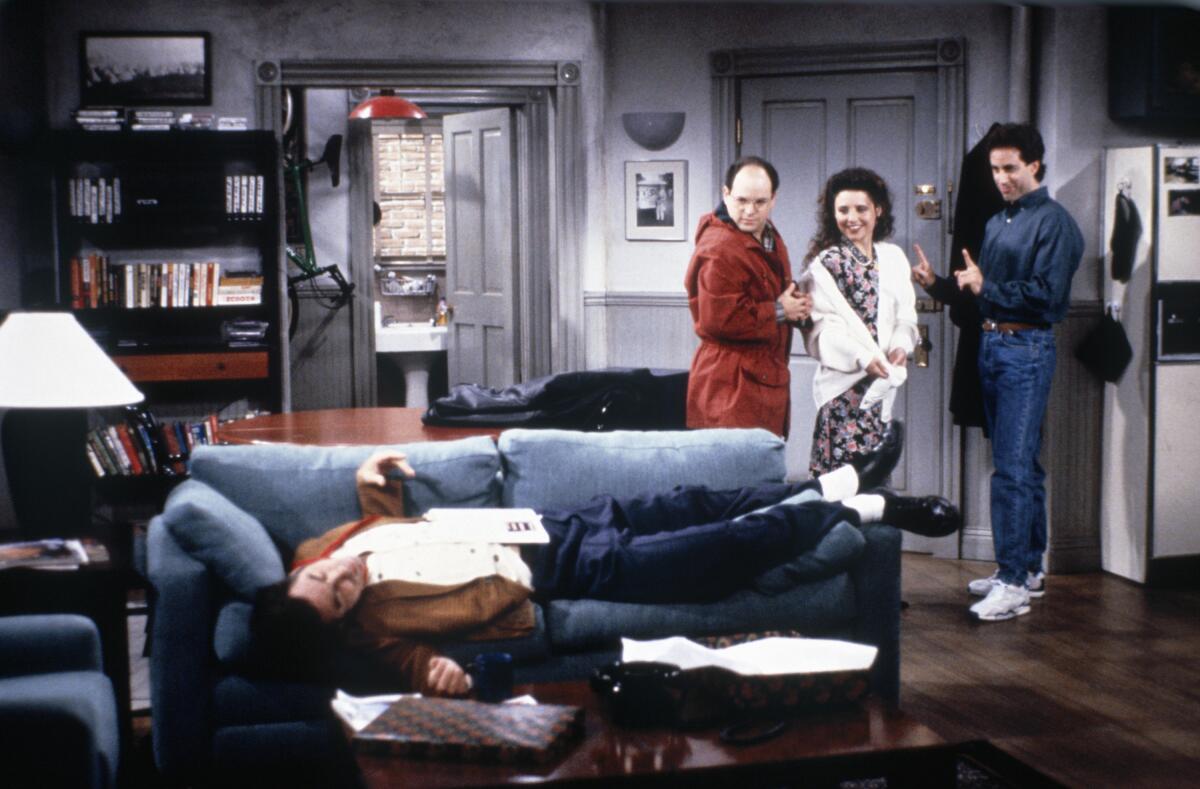 Three people stand in the doorway to an apartment and look at a man lying on a couch