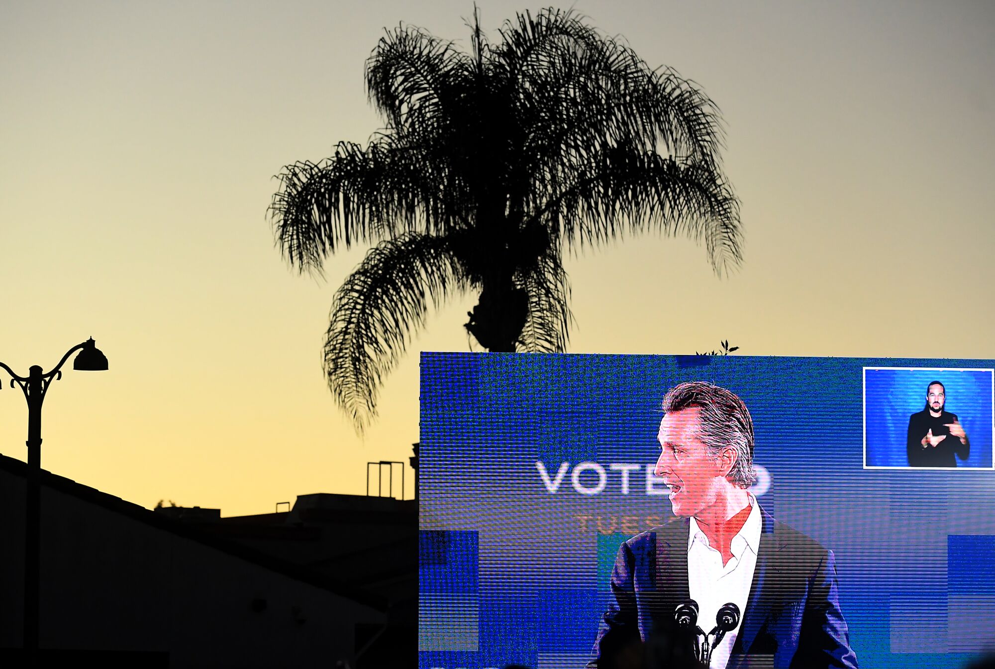 California Gov. Gavin Newsom is seen on a large outdoor monitor in the evening, with an inset image of a person signing.