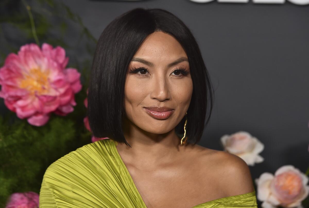 Jeannie Mai poses in a chartreuse-colored gown showing one bare shoulder against a floral backdrop