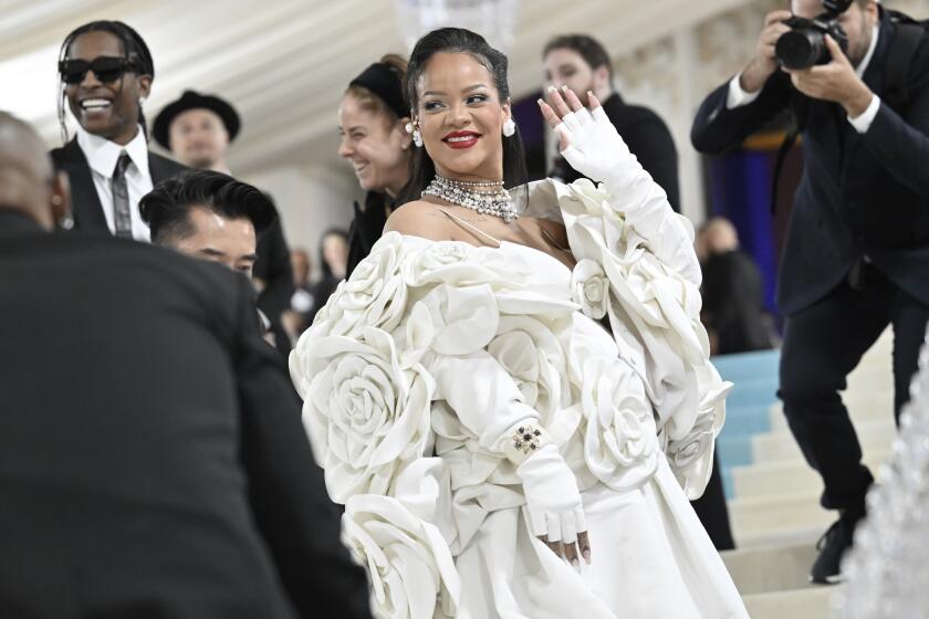 Rihanna in a white dress with large rose appliqué posing at the stairs of the Met Gala with her left hand waving