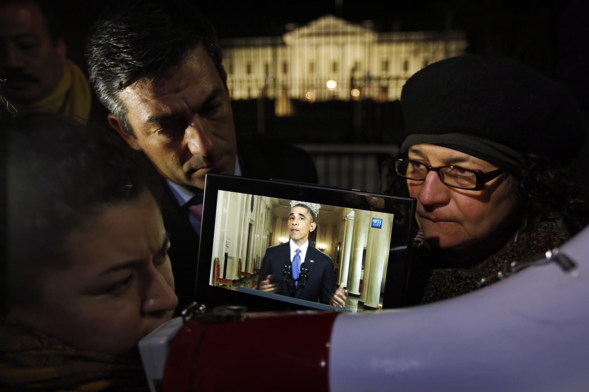 A person uses a megaphone outside the White House after dark as others watch a small screen showing President Obama speaking.