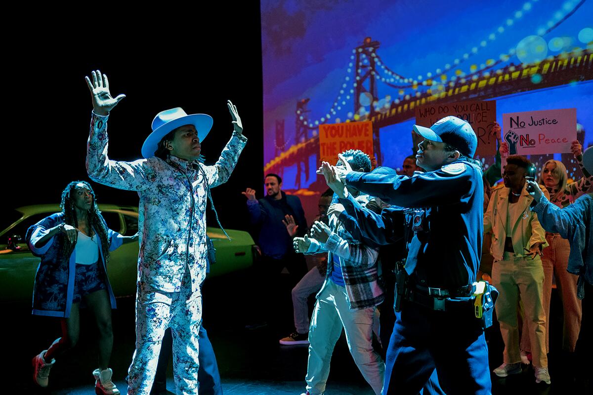 A police office points a gun at a man in a vivid suit on stage, with the Golden Gate Bridge as a backdrop