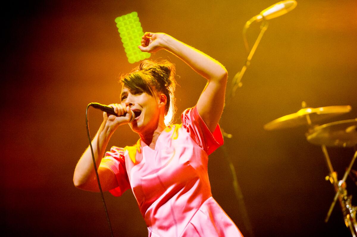 Kathleen Hanna, in a pink satin dress, sings into a mic on stage. 