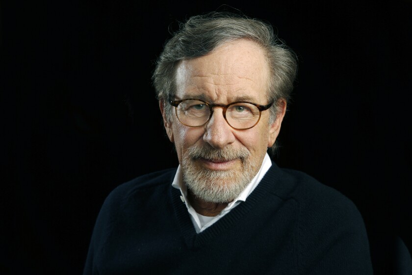 Steven Spielberg looks into the camera while wearing a sweater and glasses against a black background.
