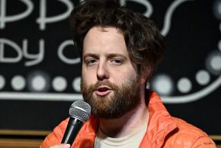 Comedian Dex Carvey is talking into a mic as he delivers a stand up set while wearing an orange puffer jacket