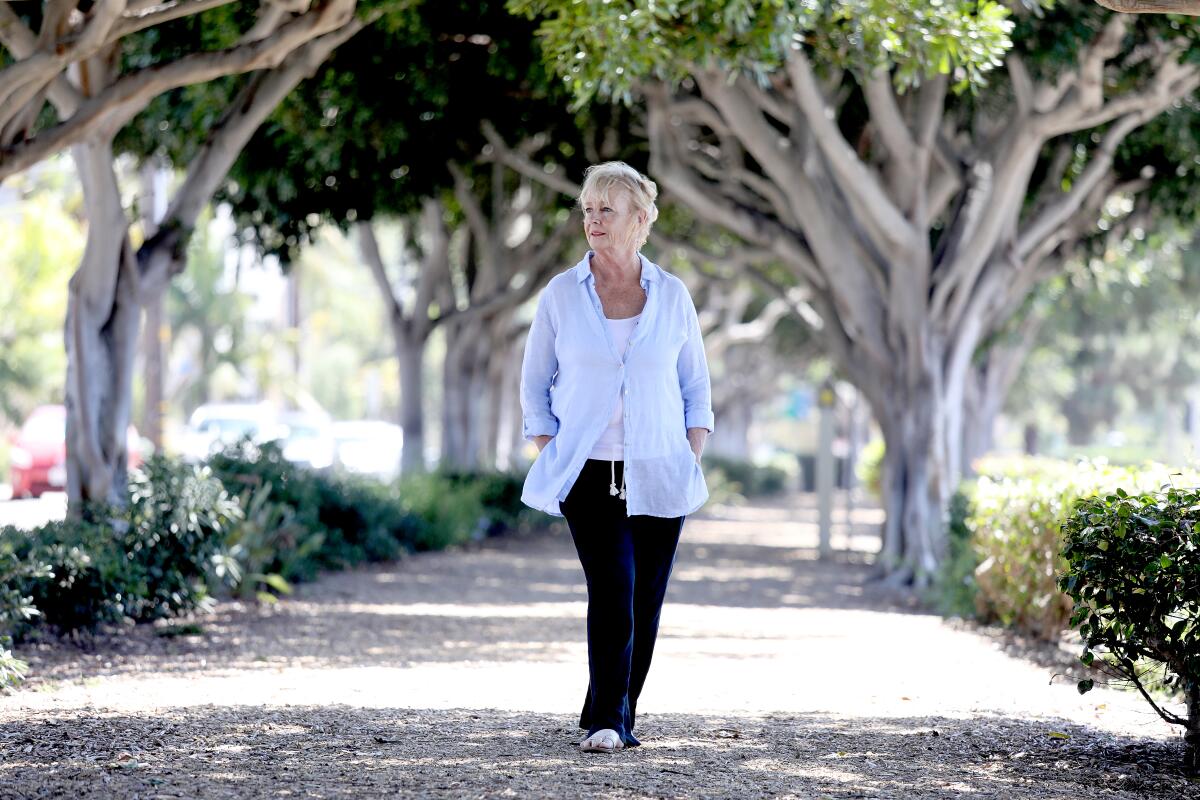 A woman with short blond hair, in a light-blue long-sleeved shirt and dark pants, walks along a path flanked by trees 