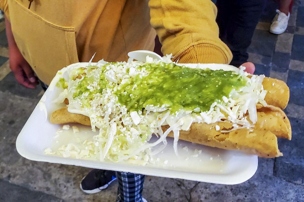 A person wearing a yellow sweatshirt holds out a foam plate of flautas covered in green sauce.