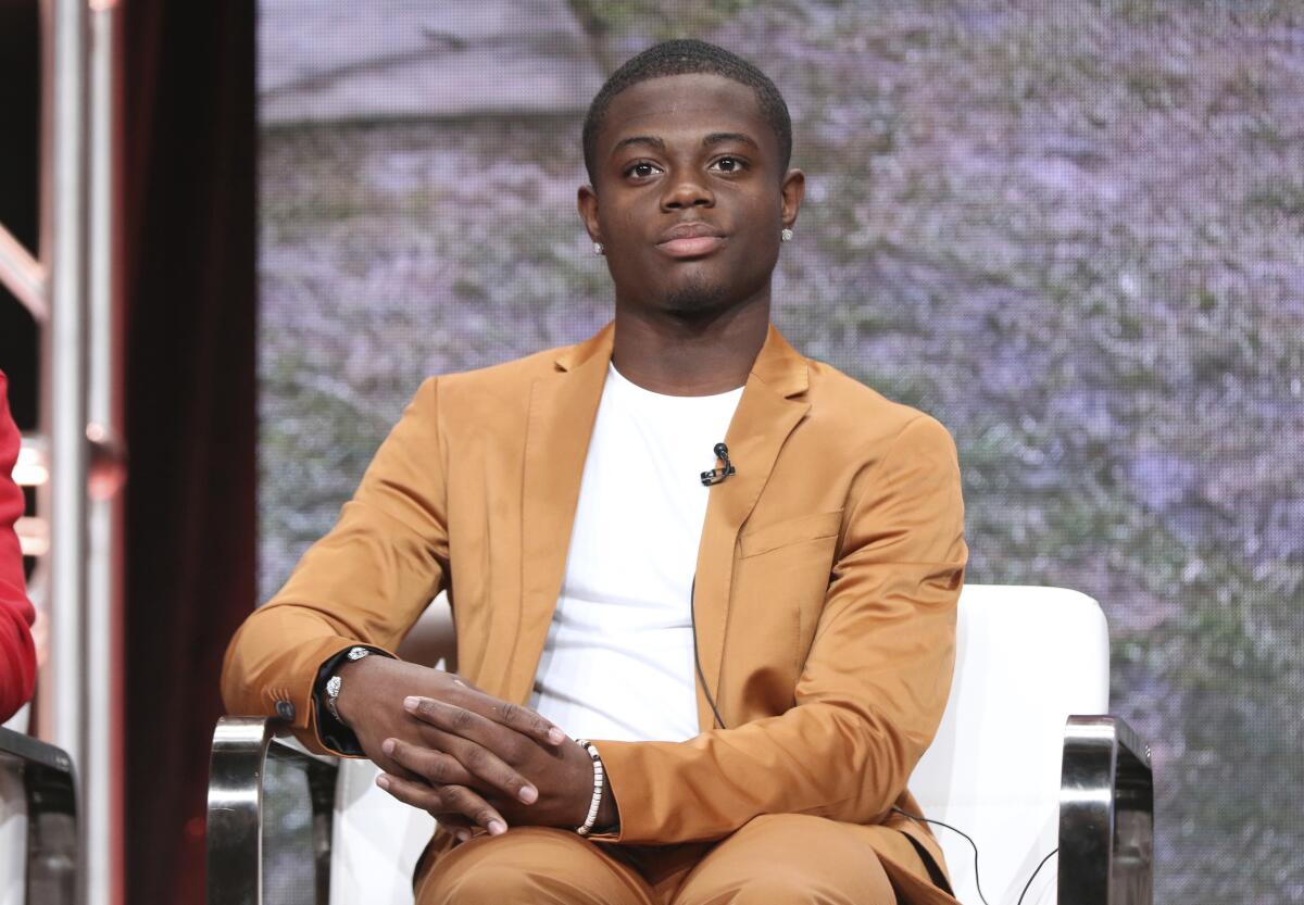 A Black man in a tan suit and a white t-shirt sitting in a white chair with his hands on the armrest