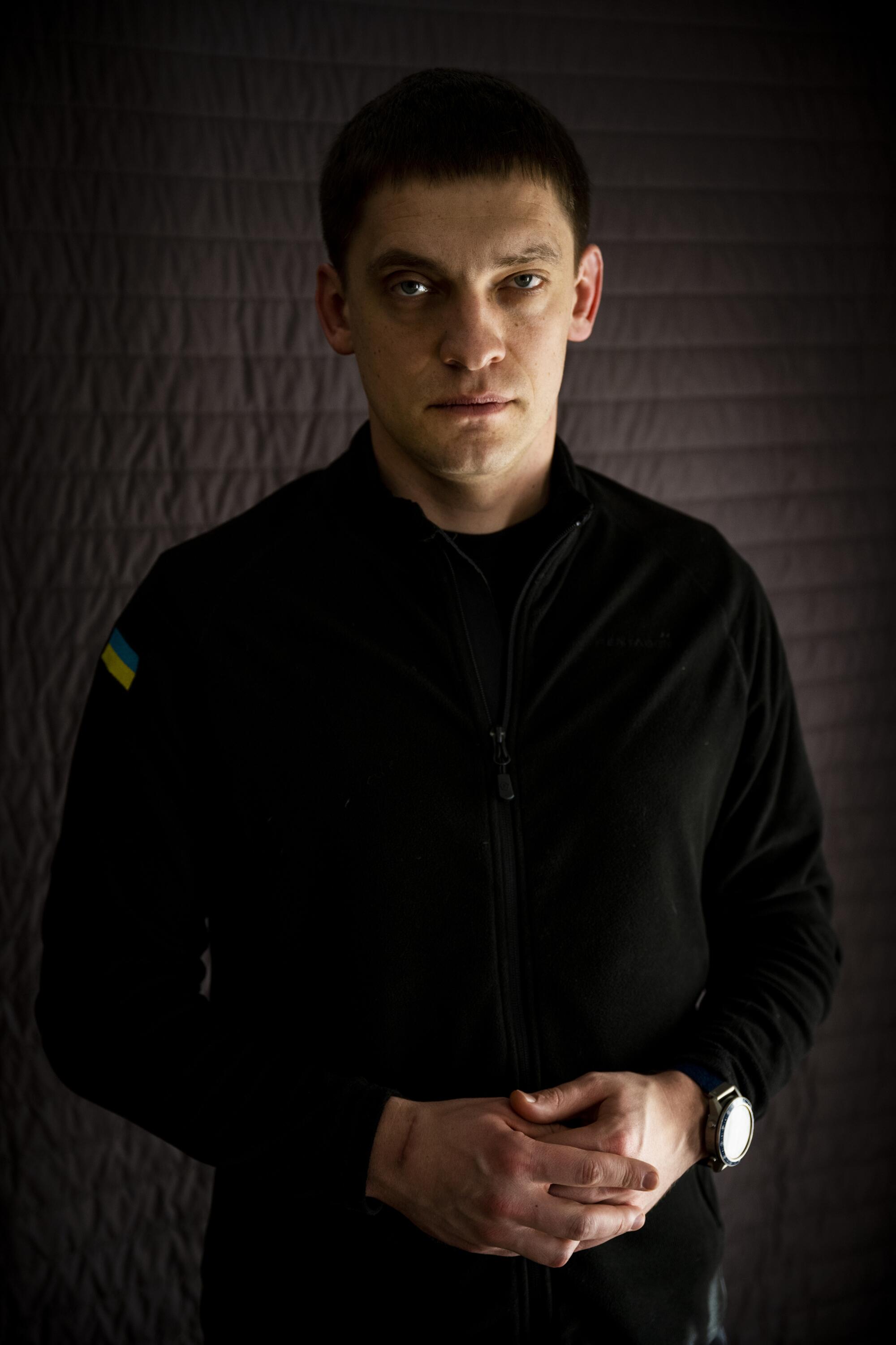 A portrait of a man in his 30s, looking right into the camera, with his hands together in front of him