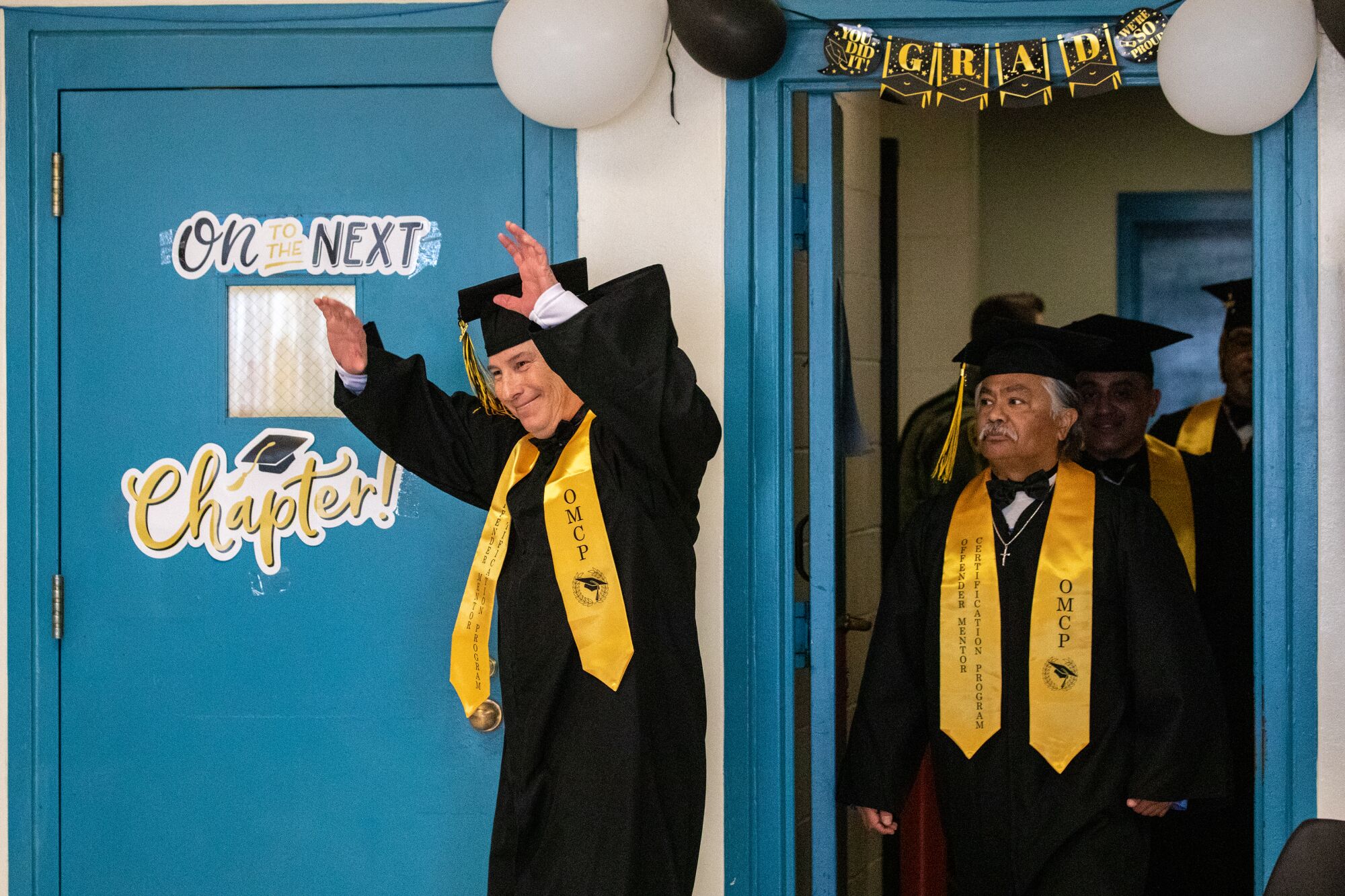 two men in cap and gowns and yellow sashes walk through a door, one with his hands in the air 