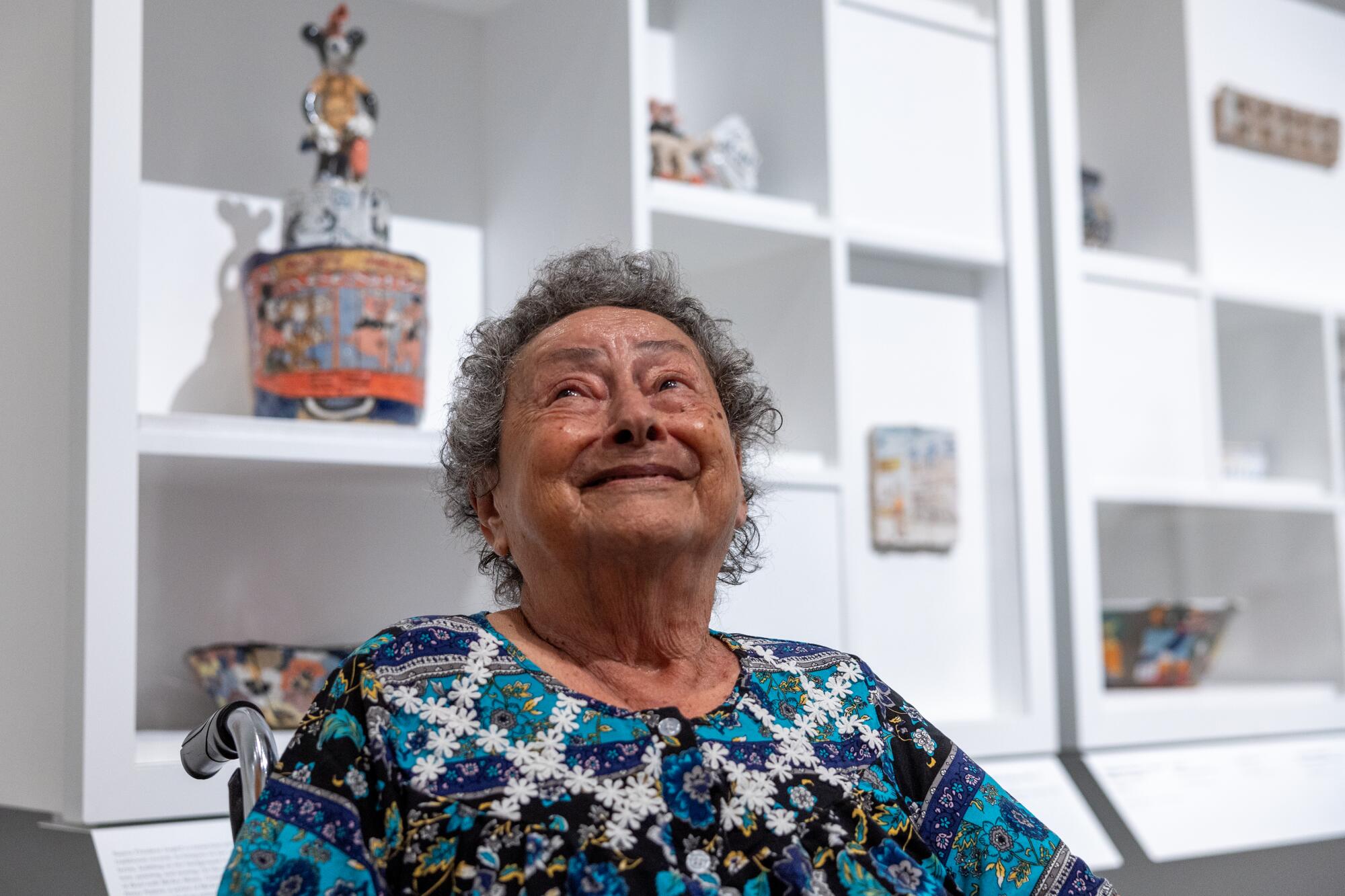 A smiling older woman in a patterned top looks up in front of shelving with artwork on them.