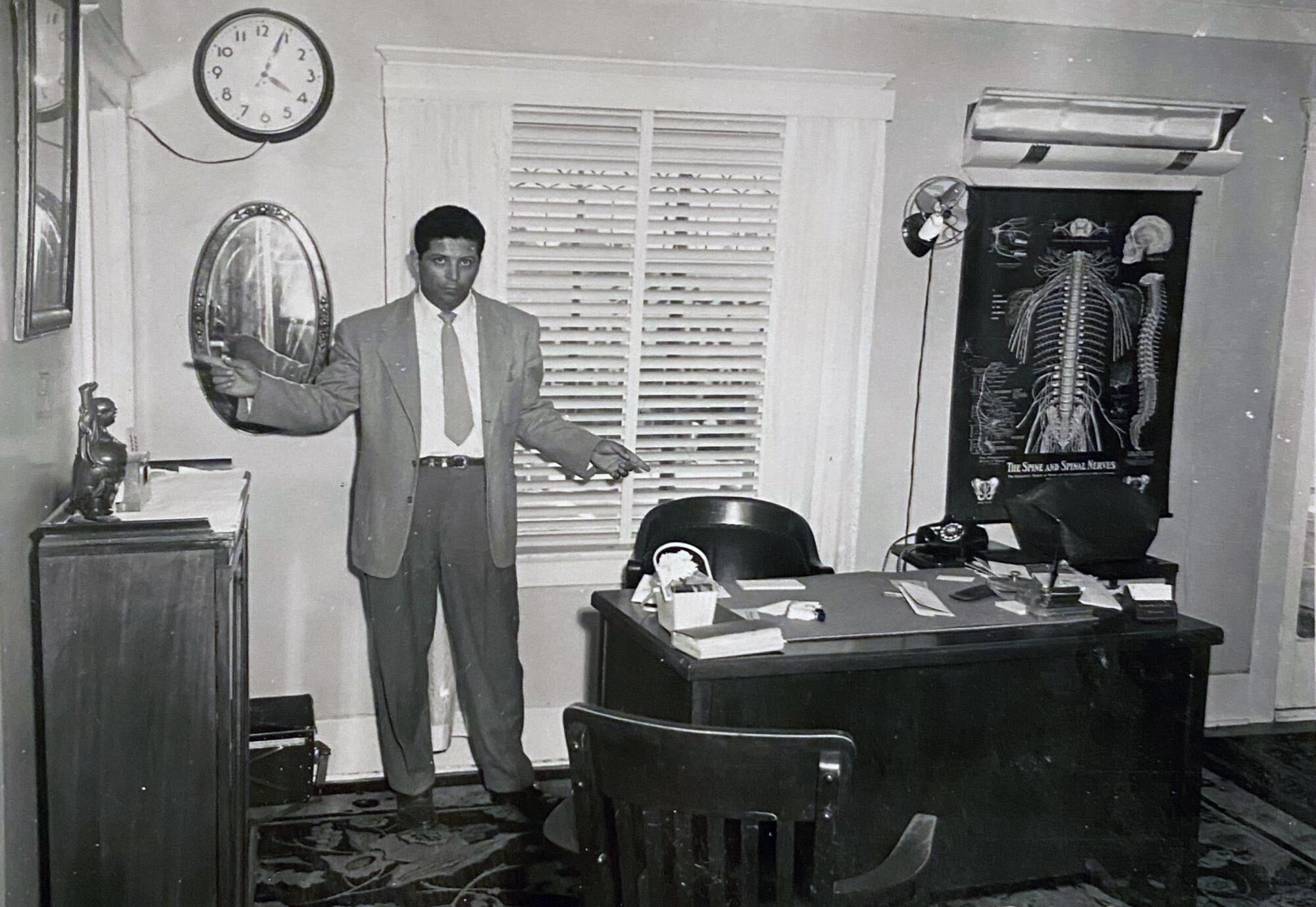 Danny Galindo wearing a 1960s-style light suit and light tie standing beside a desk and other bedroom furniture.