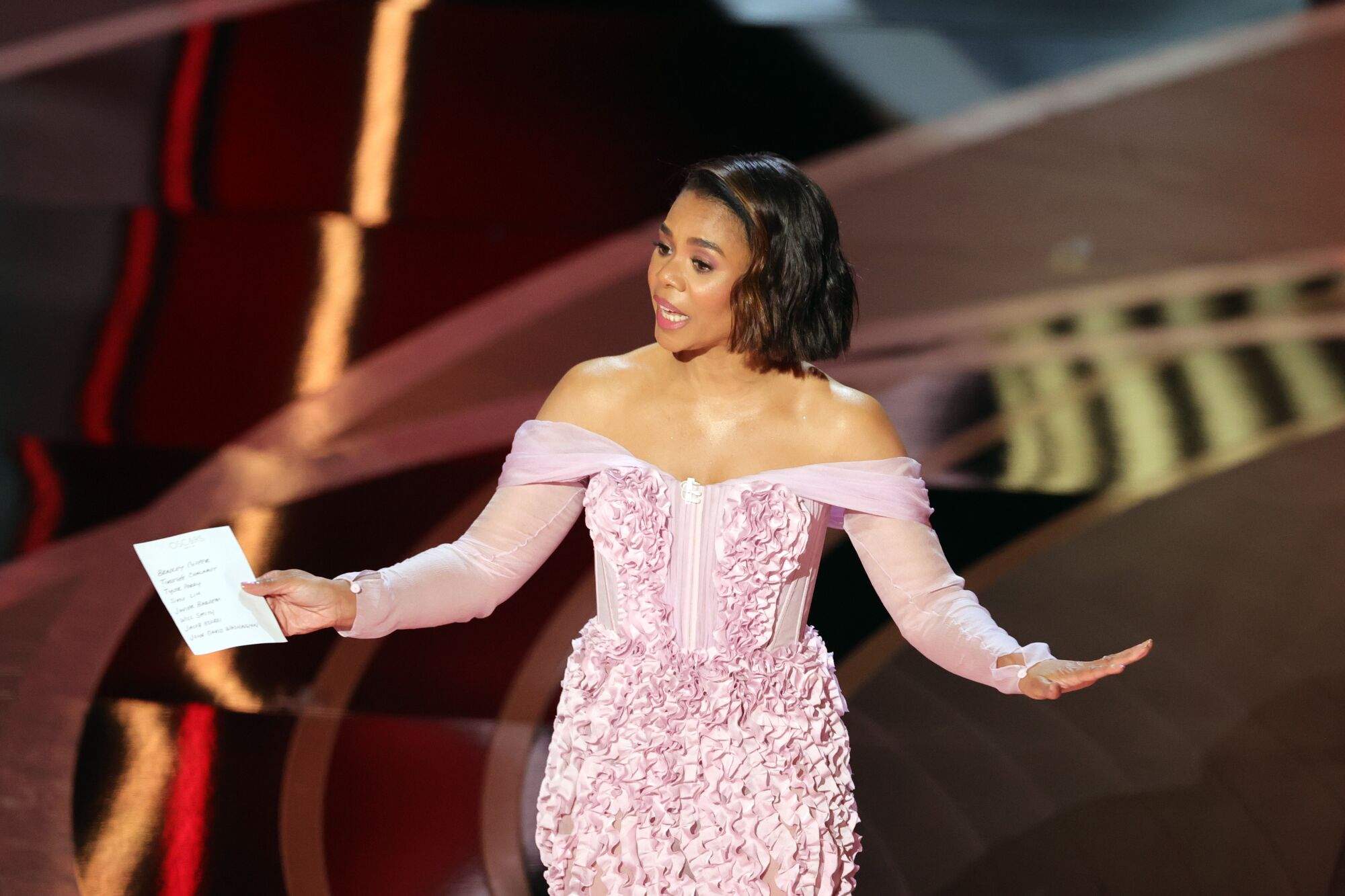 A woman in a pink textured gown speaks onstage 