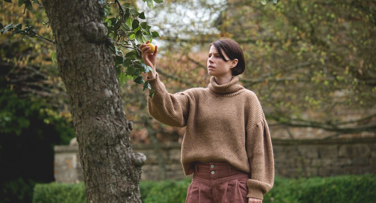 A woman in a large sweater grasps a fruit growing from a tr