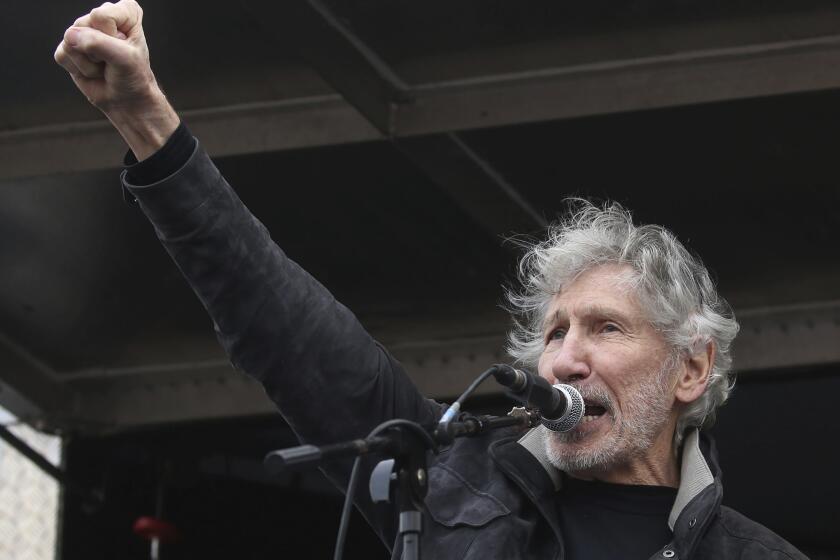 A man with gray hair and a scruffy beard speaks at a microphone while raising his right hand in a fist