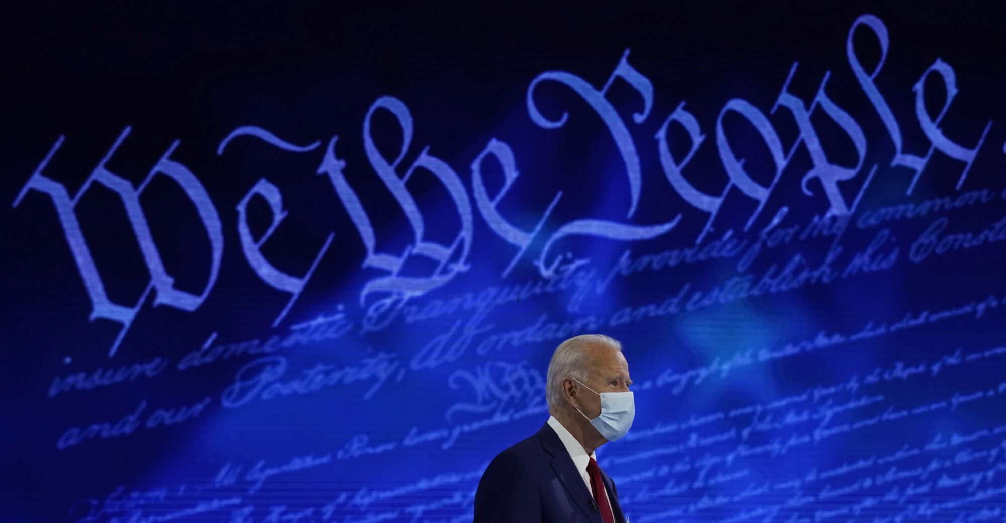 Joe Biden in front of a blue wall with the words We the People