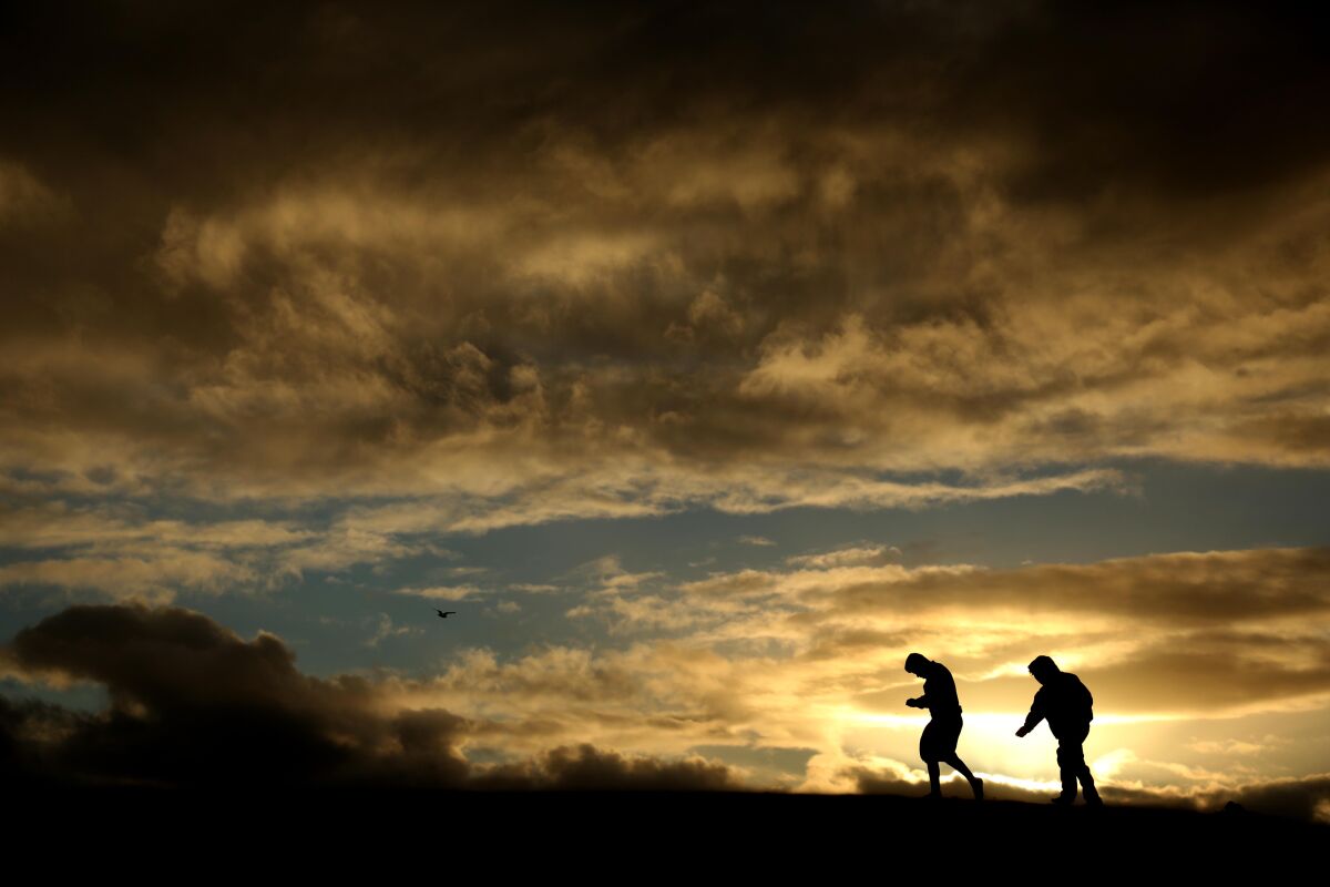 Two boys are silhouetted against the sun setting in a cloudy sky.