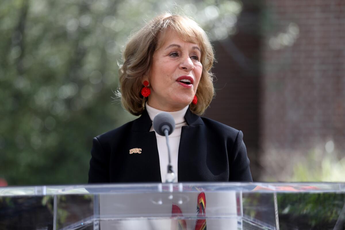 USC president Carol Folt stands at a podium and addresses a crowd while standing in a rock garden on campus. 