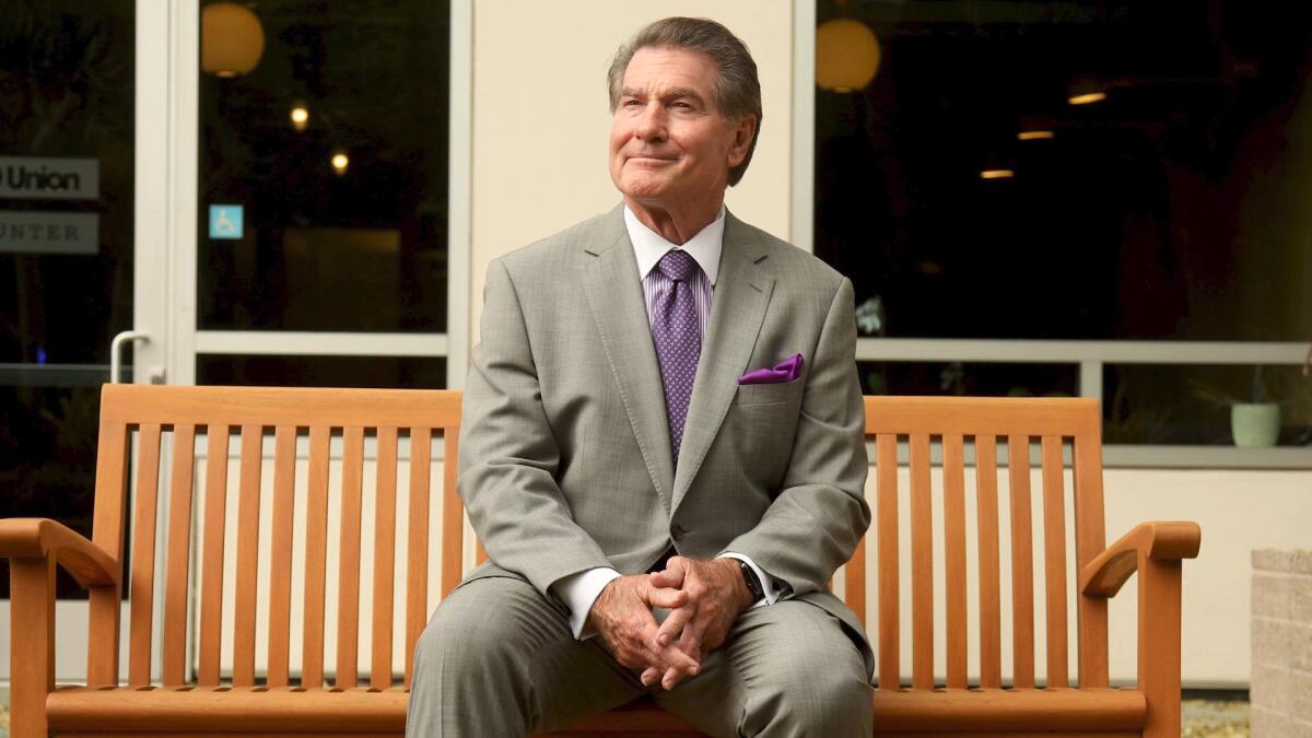 A man in a gray suit and purple tie smiles while sitting on a wooden bench