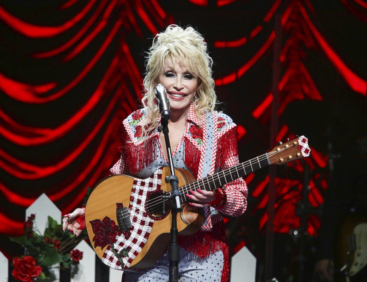 Dolly Parton wearing a red fringe blouse holding a guitar and standing in front of a microphone on a stage