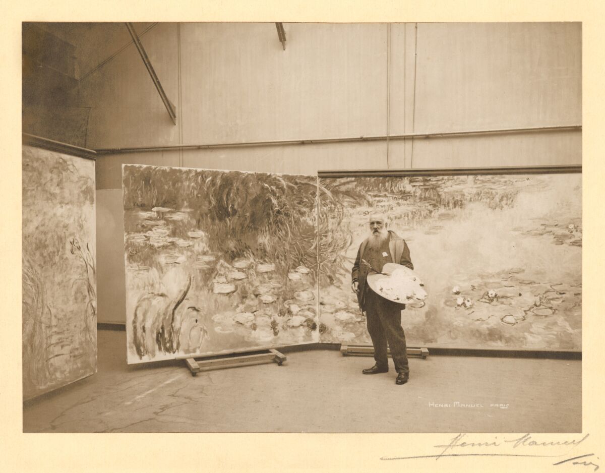 An old man  with a long beard poses in front of a large painting while holding a painter's palette. 