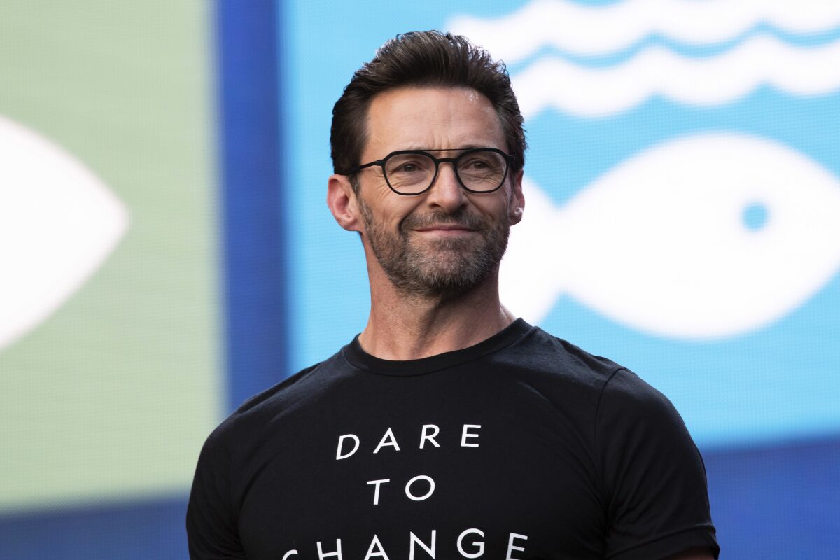 Hugh Jackman smiles slightly on an event stage wearing a T-shirt that says "Dare to Change."