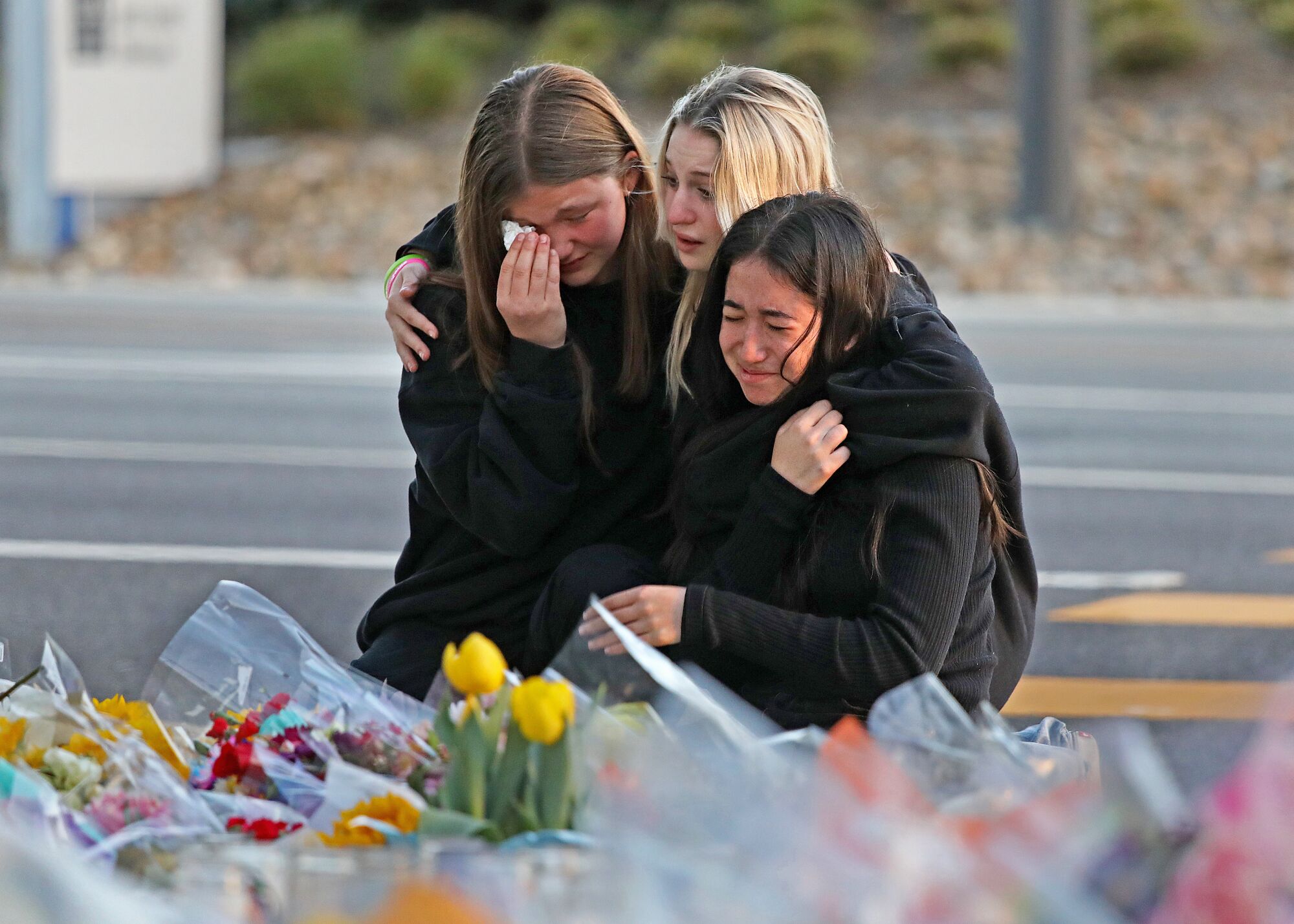 Три девочки-подростка обнимаются и плачут рядом с придорожным мемориалом из цветов.