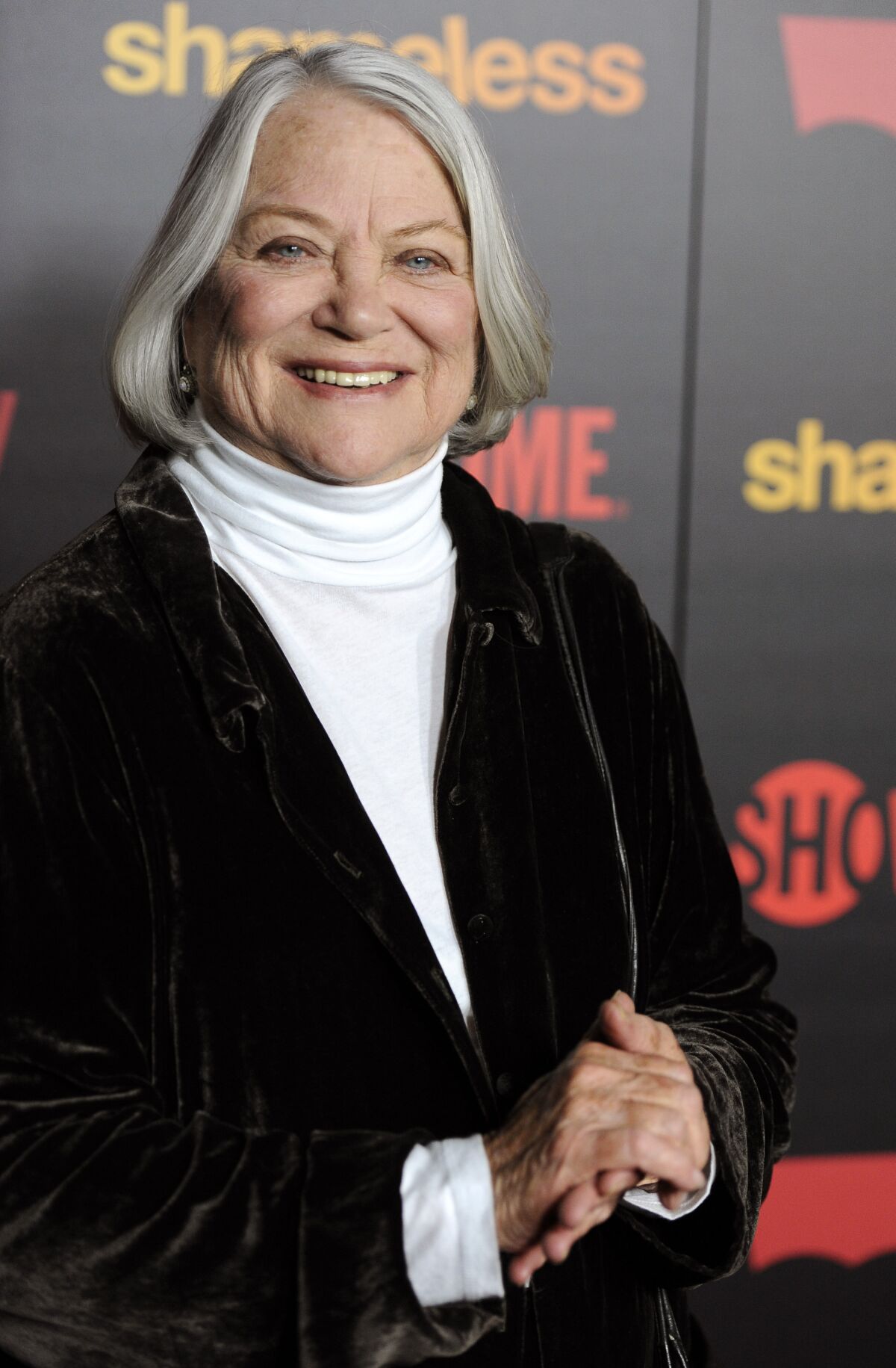 A woman with a gray bob, wearing a white turtleneck and a black jacket smiles