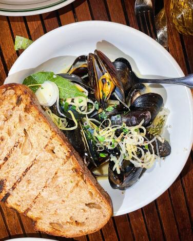 An overhead vertical photo of a bowl of mussels from L&E Oyster Bar in Silver Lake. A piece of grilled bread on rim of bowl.