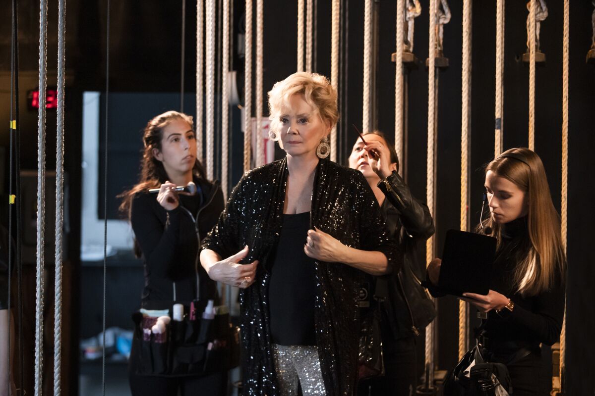 Three women with makeup and hair tools stand around Jean Smart, in a sparkly black jacket, backstage.