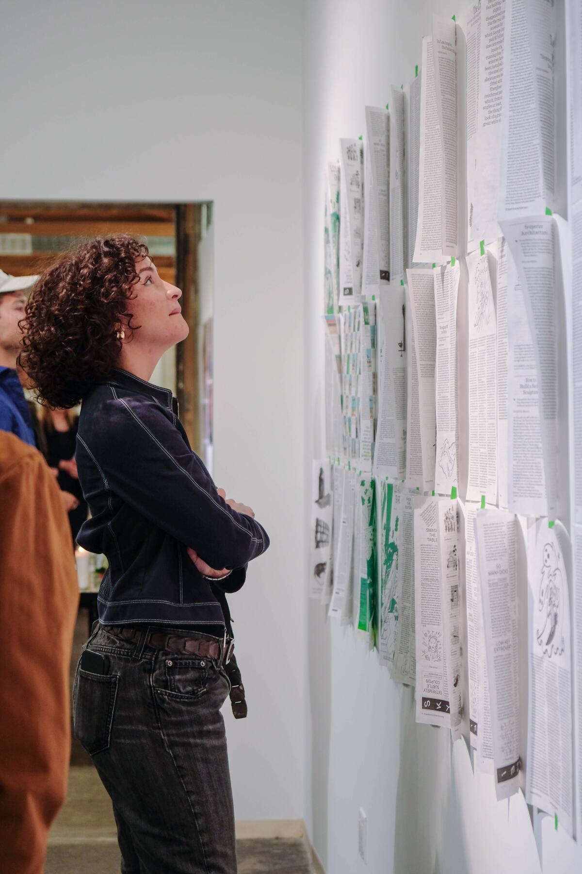 A woman with curly hair looks at a series of magazine spreads pinned to a wall inside a gallery.