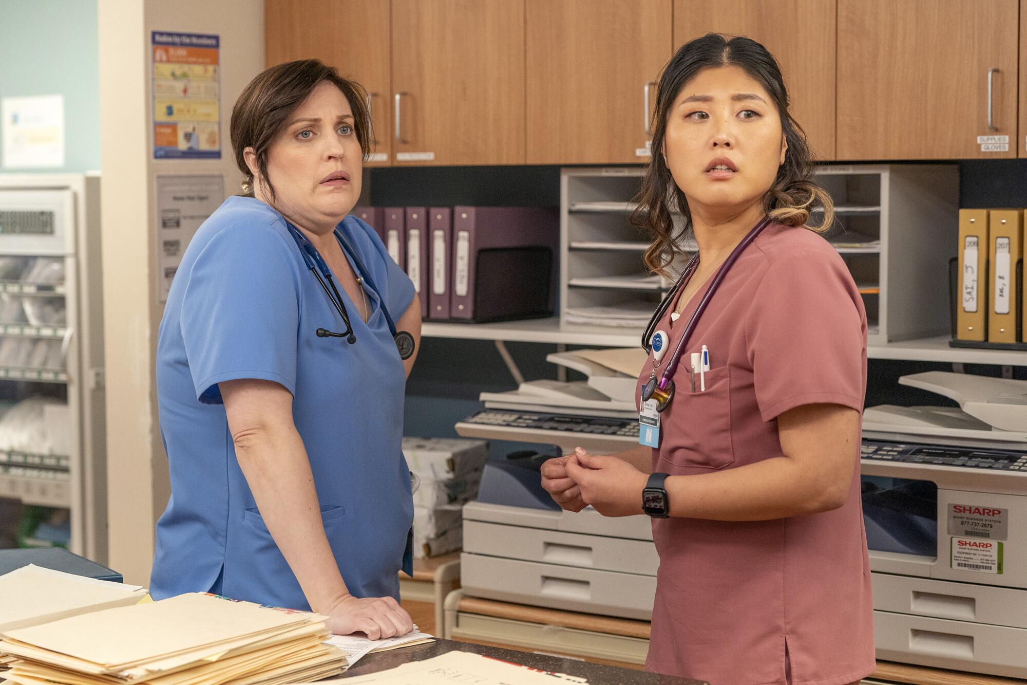 Two nurses in scrubs stand among office shelving filled with binders and files.