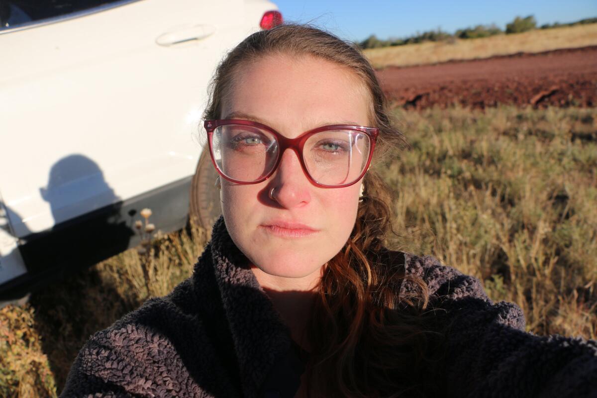 A woman in glasses takes a selfie next to a car in a field