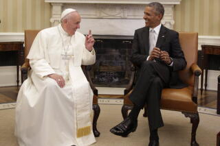 A man in white skullcap and religious vestments smiles as he speaks to another man in a dark suit, also seated