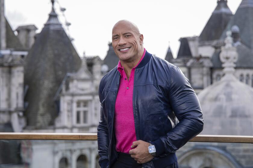 Dwayne Johnson in a hot pink shirt and a leather jacket posting in front of buildings in London