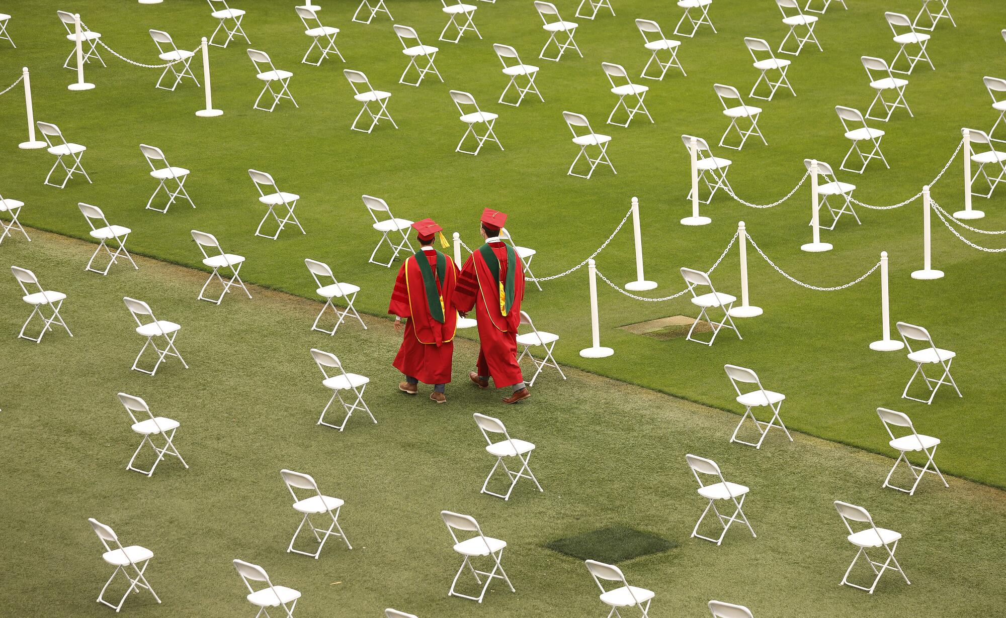 Two students in red graduation regalia walk through a grid of white folding chairs on a field