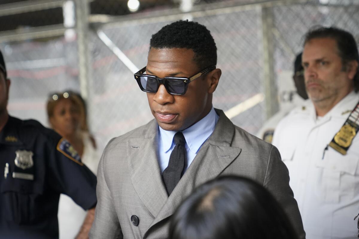 Jonathan Majors leaves a courthouse while wearing a gray jacket over a blue shirt with a black tie and sunglasses